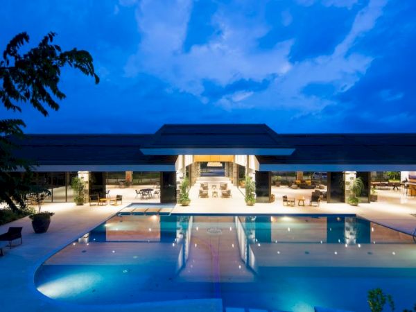 A luxurious pool area is lit up at night, with a modern building and outdoor seating in the background, under a partly cloudy sky.