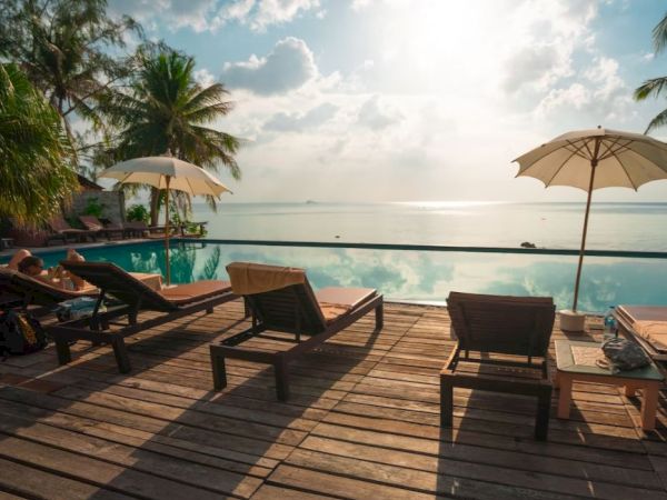 A serene poolside area with lounge chairs, umbrellas, and a stunning view of the ocean under a partly cloudy sky.
