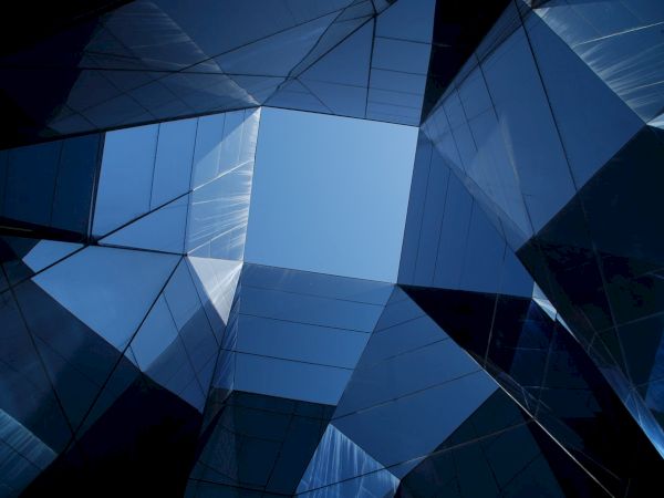 The image shows a geometric arrangement of mirrored glass panels forming a square opening to a clear blue sky above.