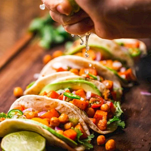 The image shows several tacos filled with chickpeas and vegetables, with a hand squeezing lime juice over them.
