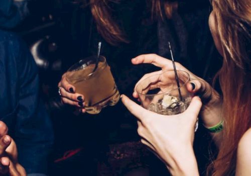 People are at a party or club, holding drinks and socializing. The focus is on their hands, drinks, and laughter, creating a lively atmosphere.