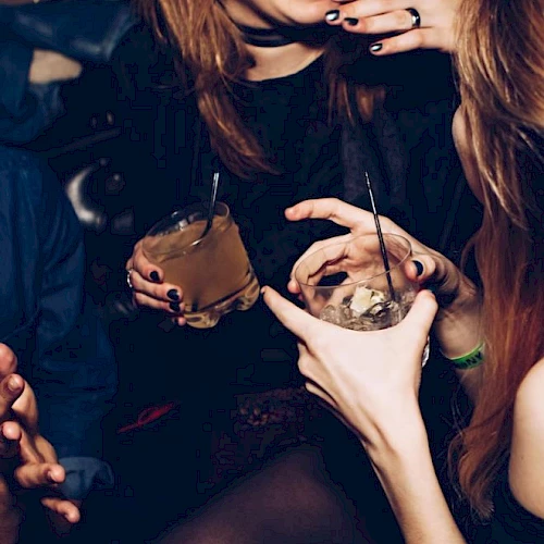 People are at a party or club, holding drinks and socializing. The focus is on their hands, drinks, and laughter, creating a lively atmosphere.