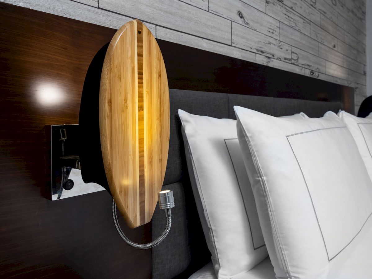 A modern bedroom features white pillows and wooden wall-mounted lamps shaped like surfboards with a rustic wood accent wall.