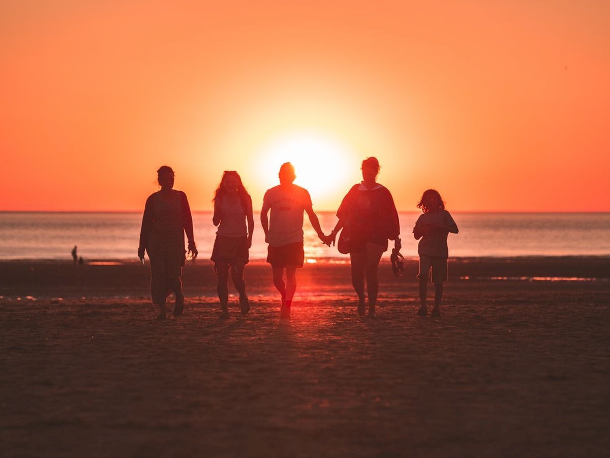 A group of five people walk hand in hand on a beach at sunset, silhouetted against the glowing orange sky with the sun low over the horizon.