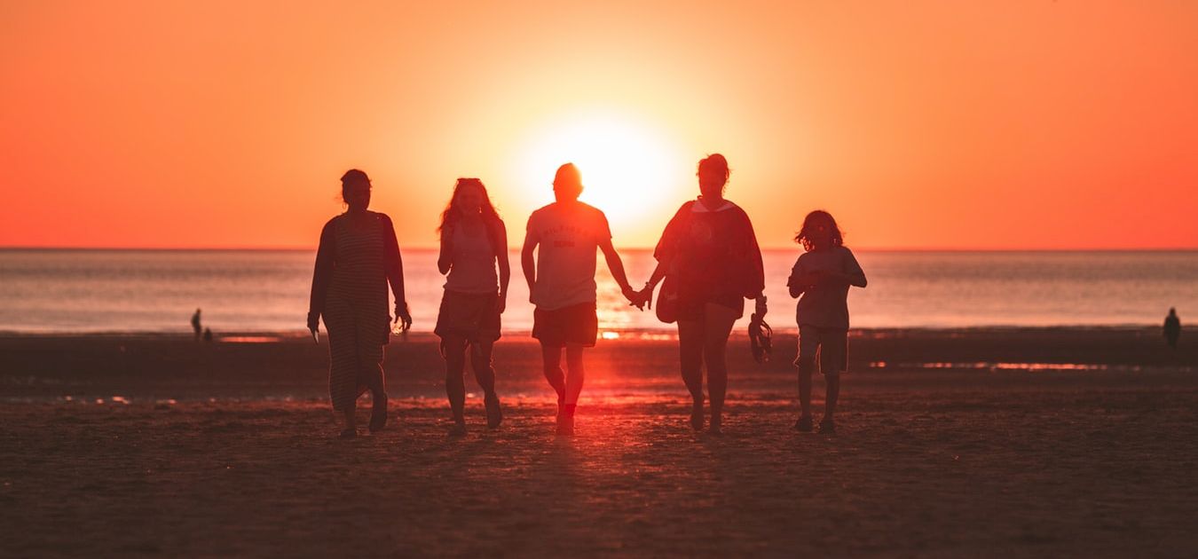 A group of five people walk hand in hand on a beach at sunset, silhouetted against the glowing orange sky with the sun low over the horizon.