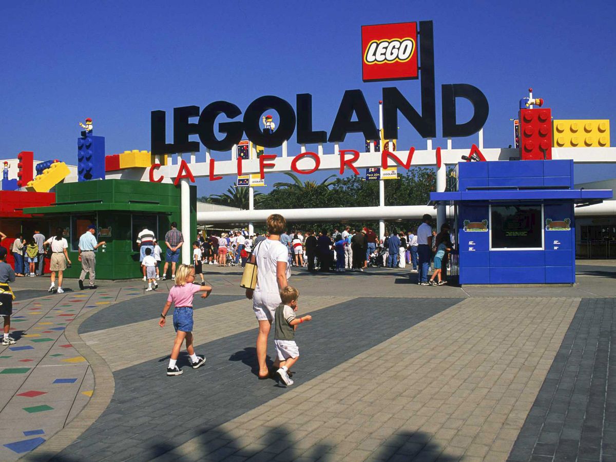 The image shows the entrance to LEGOLAND California, with people walking in the foreground and a sign with LEGO bricks in the background.