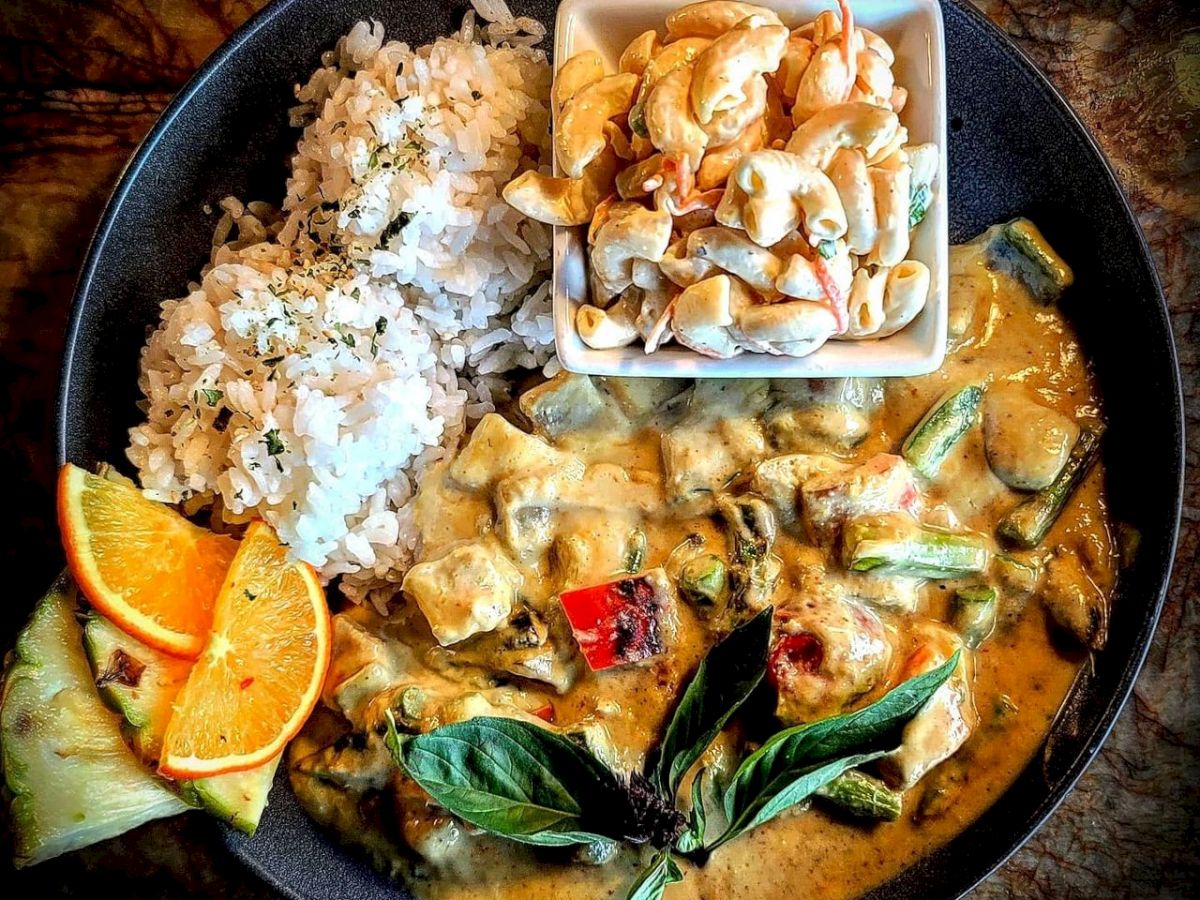 The image shows a plate with rice, a creamy vegetable curry, garnished with basil, orange slices, and a small bowl of cashews.