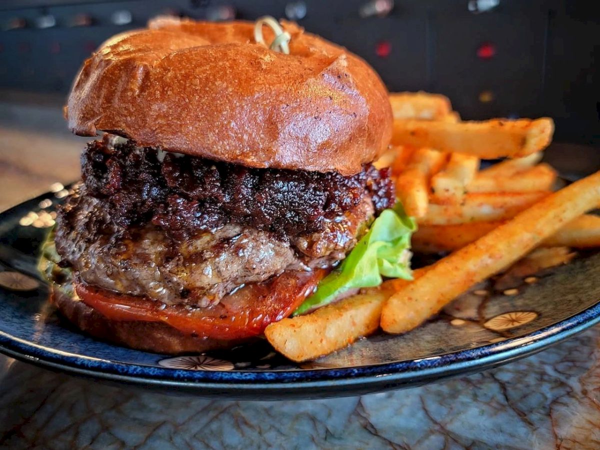 A juicy burger with lettuce, tomato, and a sesame bun, served on a blue plate with a side of golden French fries, sits on a table.