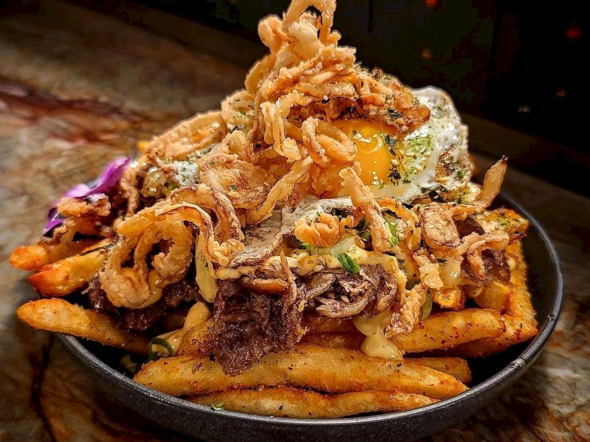 The image shows a dish of loaded fries topped with pulled meat, a fried egg, crispy onion strings, and garnished with herbs, all served in a bowl.