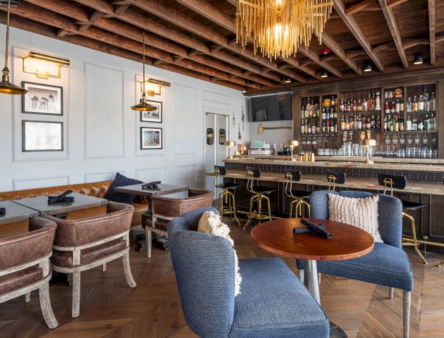 A cozy bar with a wooden ceiling, stylish lighting, a stocked bar, and comfortable seating, featuring a mix of tables, chairs, and barstools.