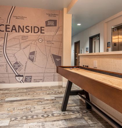 The image shows an indoor space with a large wall map of Oceanside, a desk area, a shuffleboard table, and an elevator lobby in the background.