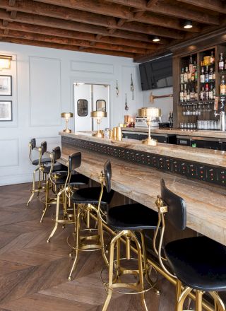 A stylish bar with a light wood counter, elegant bar stools, wall-mounted lamps, a well-stocked bar, wooden floor, and a cozy seating area.