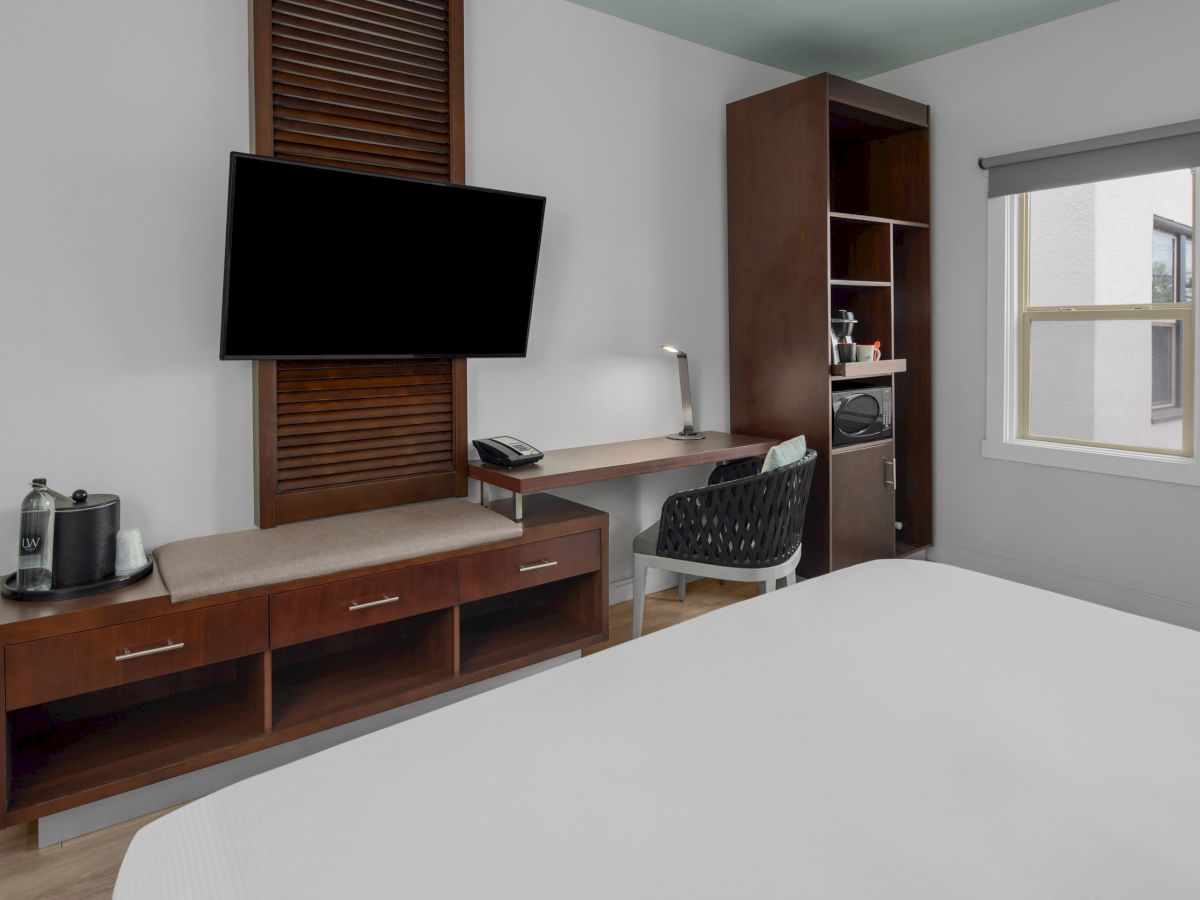 The image depicts a modern hotel room with a wall-mounted TV, desk with chair, coffee maker, open closet, and a bed, under natural light from a window.