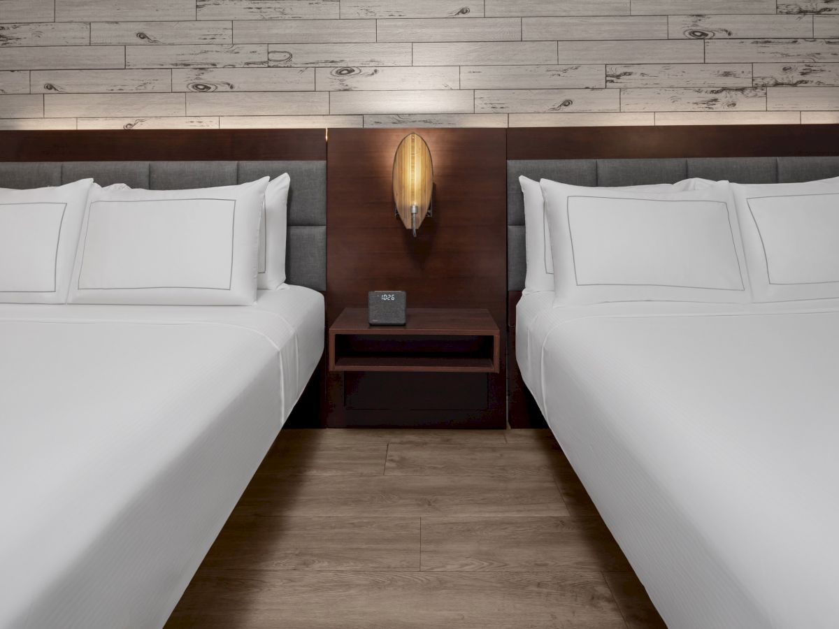 The image shows two neatly made beds in a hotel room, separated by a bedside table with a lamp and alarm clock, set against a wood-paneled wall.