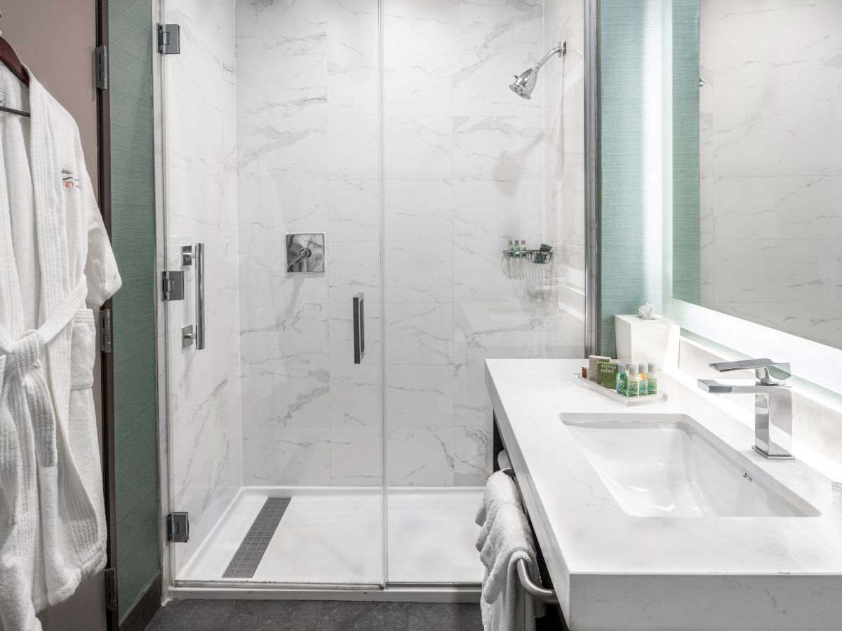 A modern bathroom with a glass-enclosed shower, white sink, toiletries, a robe hanging on a wall hook, and a large mirror with lighting.