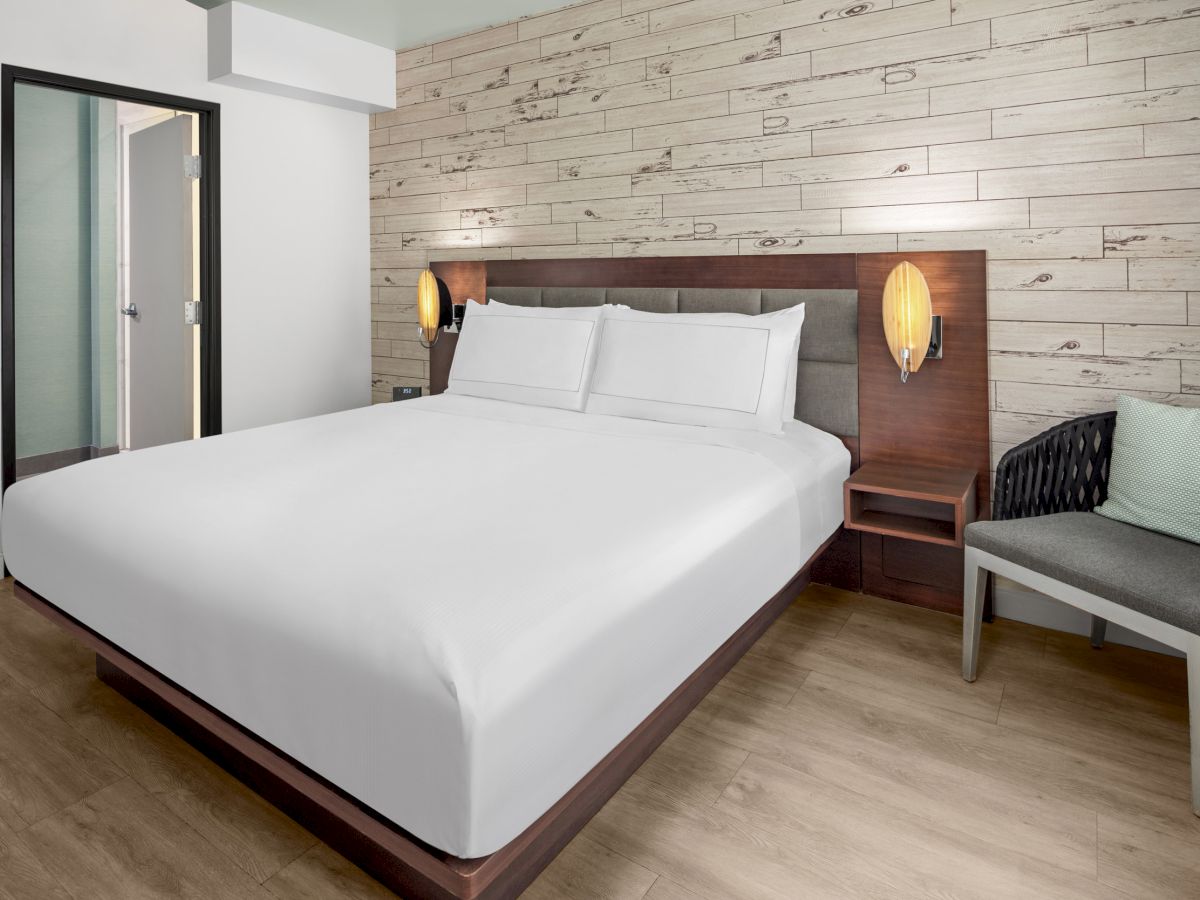 A modern hotel room with a large bed, wooden accent wall, two wall-mounted lamps, a chair with a cushion, and wooden flooring.