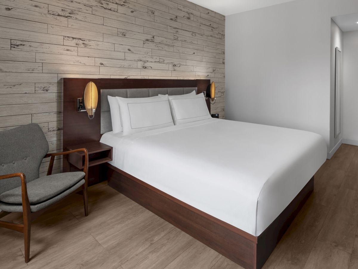 A modern hotel room with a large bed, wooden accent wall, bedside lamps, a grey armchair, wooden floor, and bright lighting.