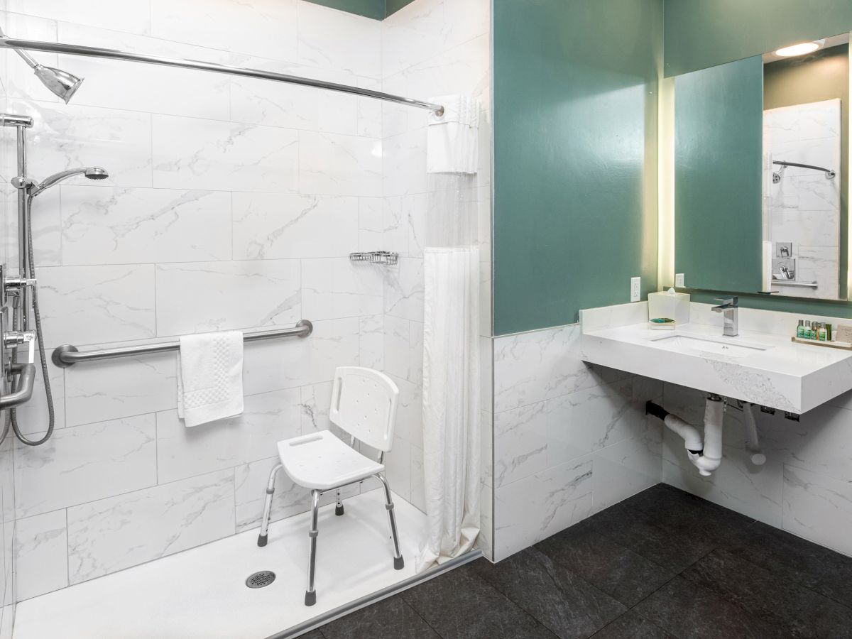 The image shows a bathroom with a walk-in shower equipped with a shower chair, grab bars, and a vanity with a large mirror and sink.