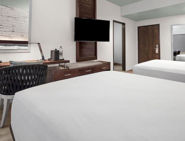 The image shows a modern hotel room with two beds, a desk, a chair, a flat-screen TV, a mirror, a phone, and some decor on the walls.