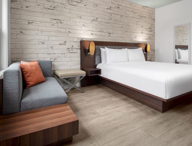 A modern hotel room with a neatly made bed, a small sofa with an orange cushion, a bench, a mirror, and light wooden flooring.