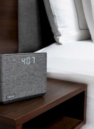 A digital clock displaying 4:07 is placed on a wooden bedside table next to a neatly made bed with white pillows and a wall outlet.