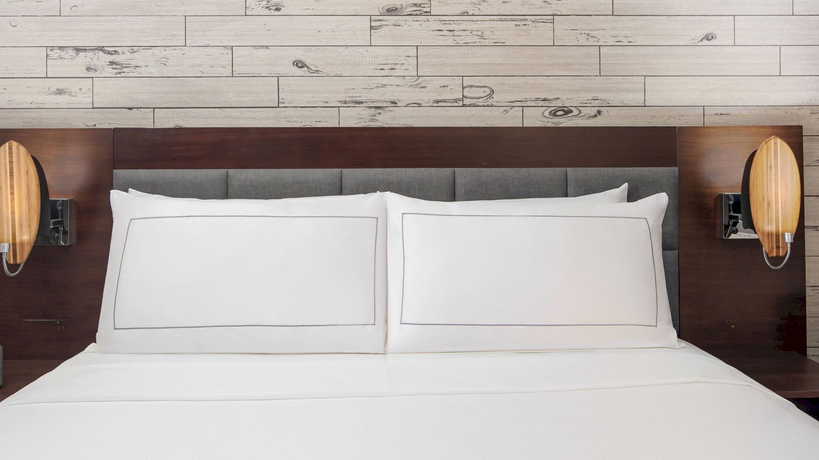 The image shows a neatly made bed with white linens, flanked by two nightstands and wall-mounted lamps, against a wooden plank accent wall.