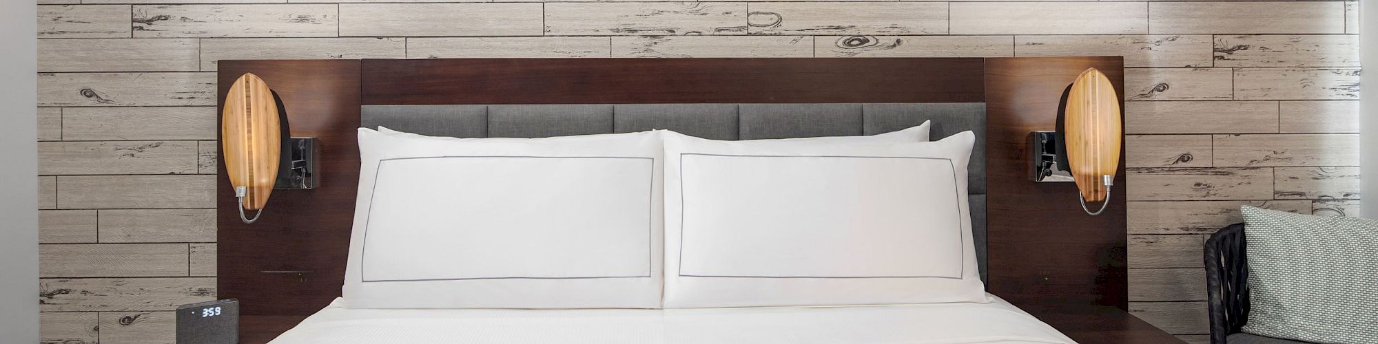 The image shows a neatly made bed with white linens, flanked by two nightstands and wall-mounted lamps, against a wooden plank accent wall.