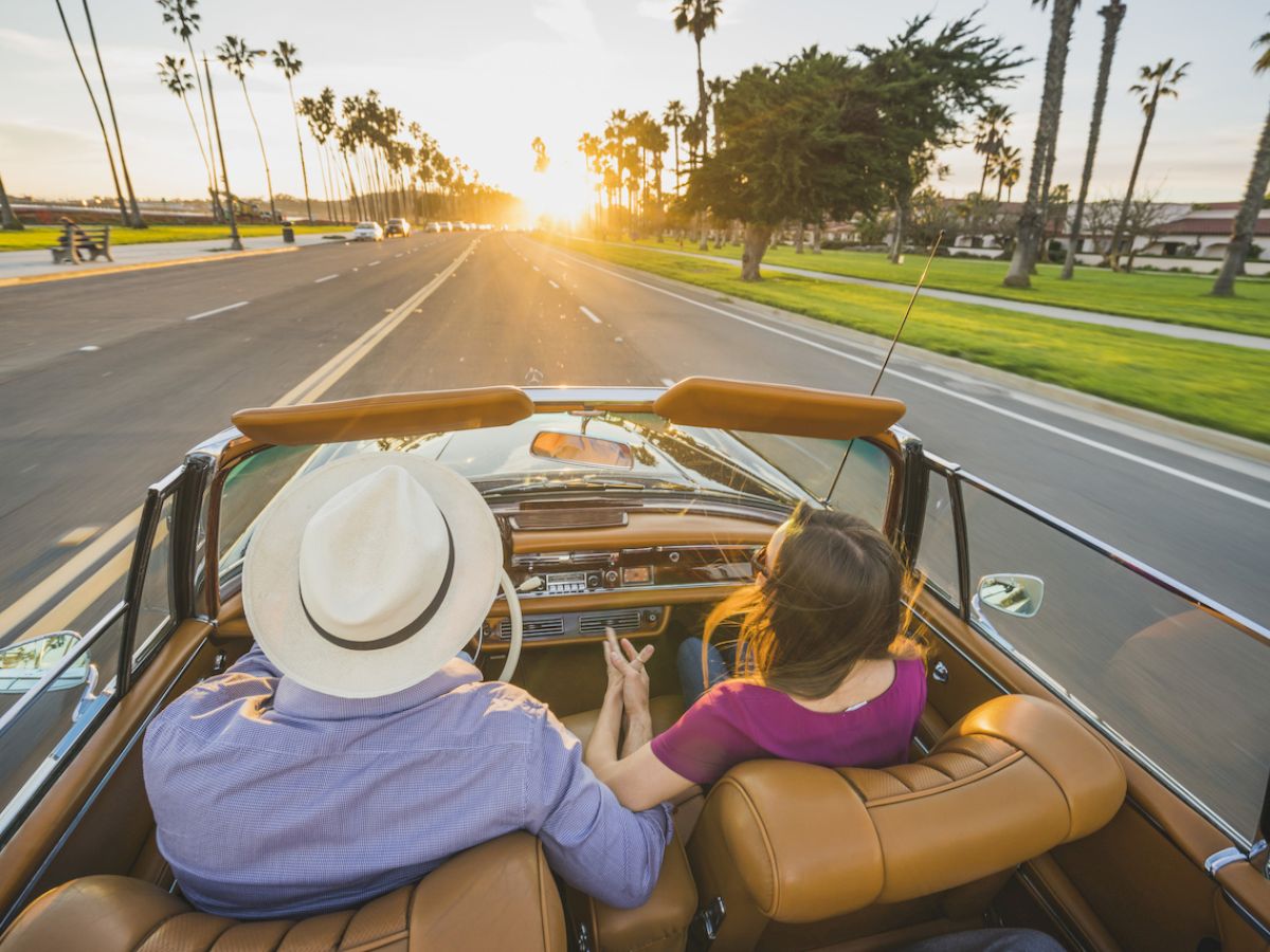 A couple is driving down a sunlit road lined with palm trees in a convertible car, enjoying a picturesque, serene evening together.