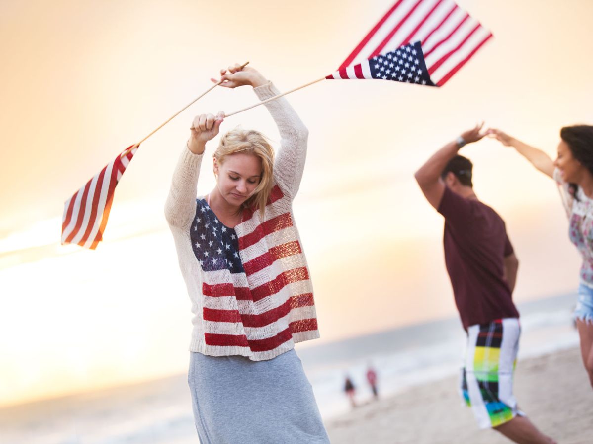 People are at the beach, waving American flags, and dancing or celebrating as the sun sets in the background.