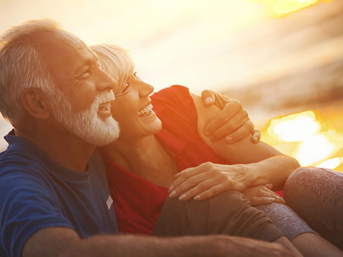 An older couple is embracing and smiling while sitting on a beach at sunset. The image captures a warm, affectionate moment.