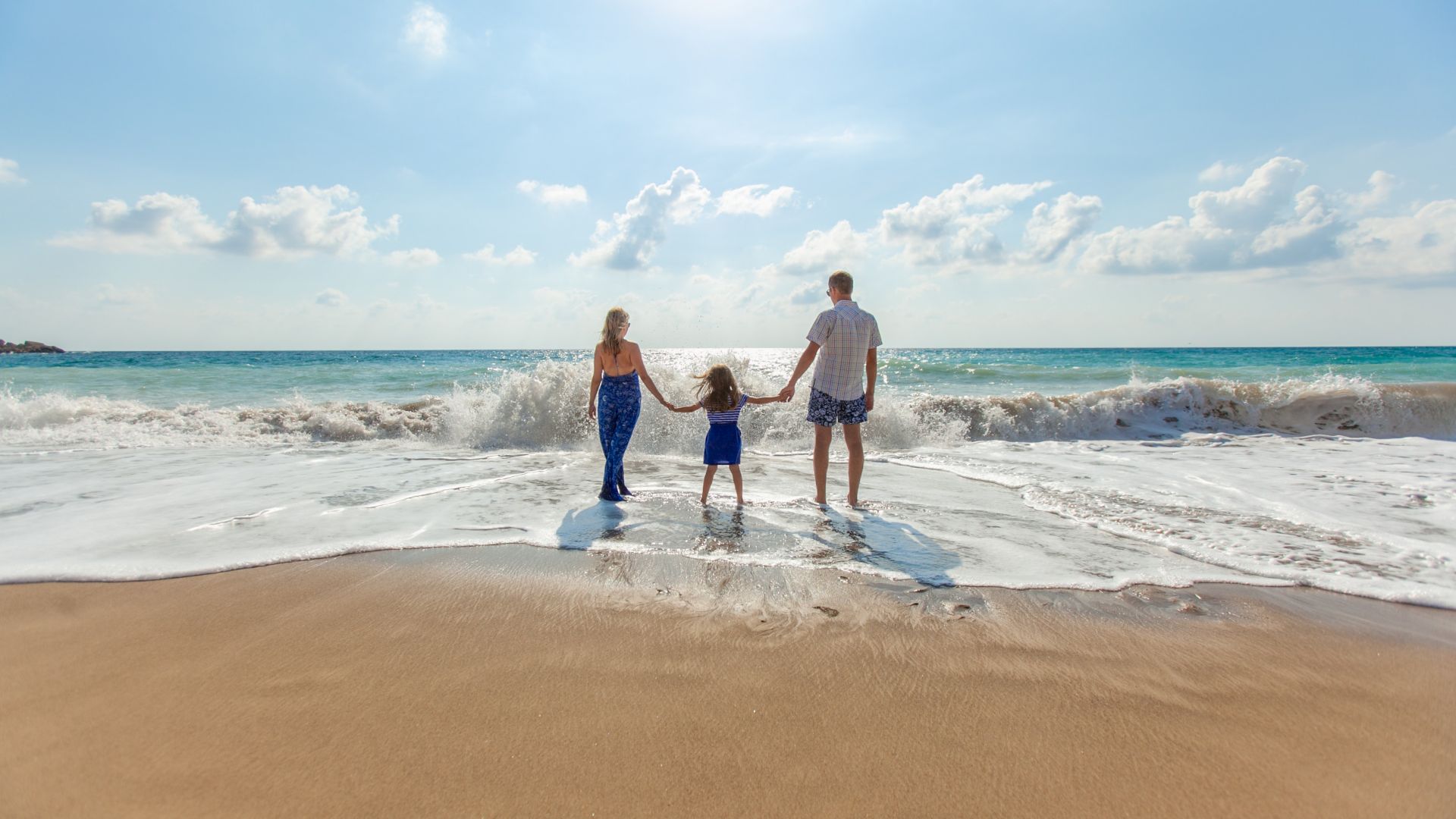 A family of three holding hands, standing at the edge of the ocean with waves crashing on a sunny day.