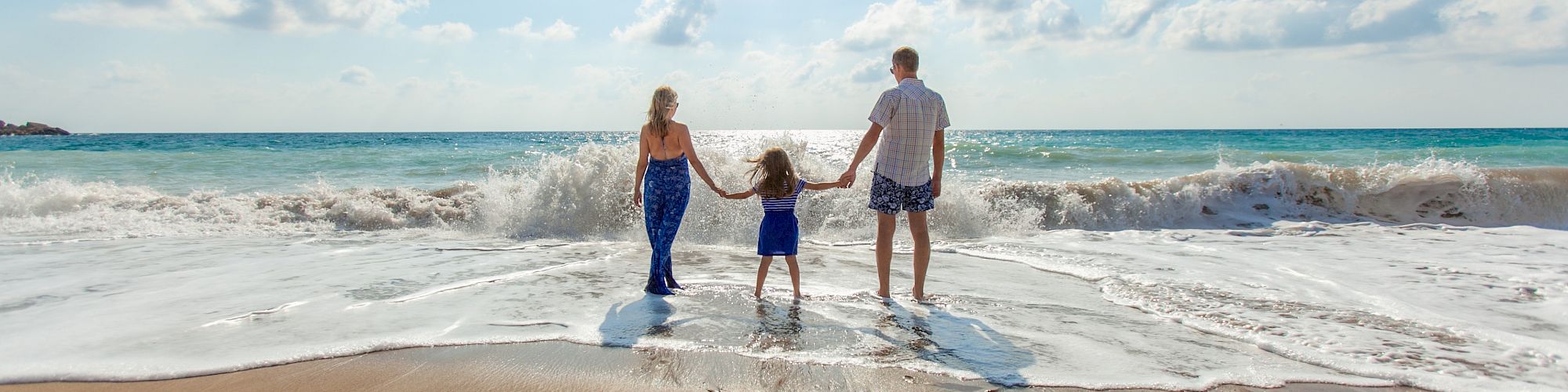 A family of three holding hands, standing at the edge of the ocean with waves crashing on a sunny day.