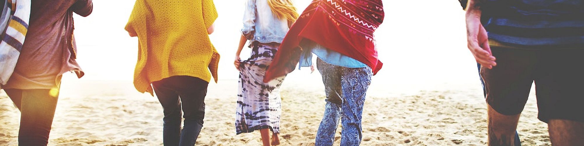 A group of people walking on the beach toward the sun, dressed casually, with one person wrapped in a blanket, enjoying a sandy outing.