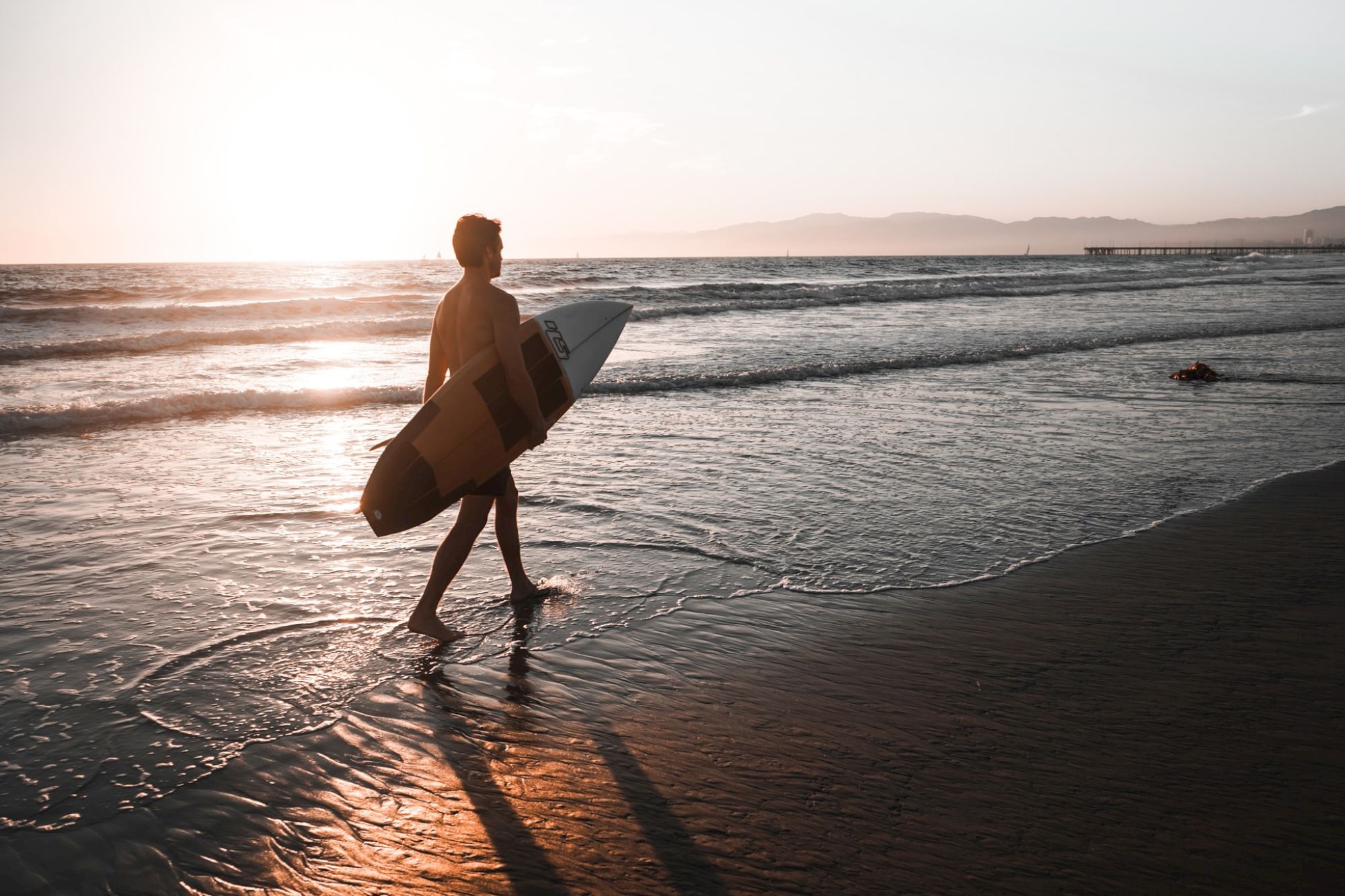 A person is walking along a beach holding a surfboard during sunset, with waves gently rolling in and mountains visible in the background.