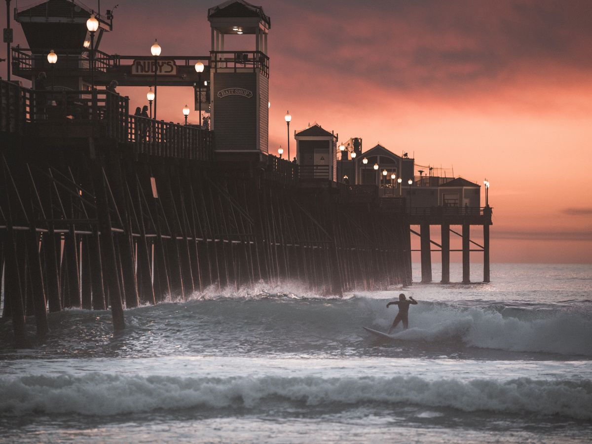 A person is surfing near a long pier with lights, against a backdrop of a stunning sunset and rolling waves, with a cloudy sky overhead.