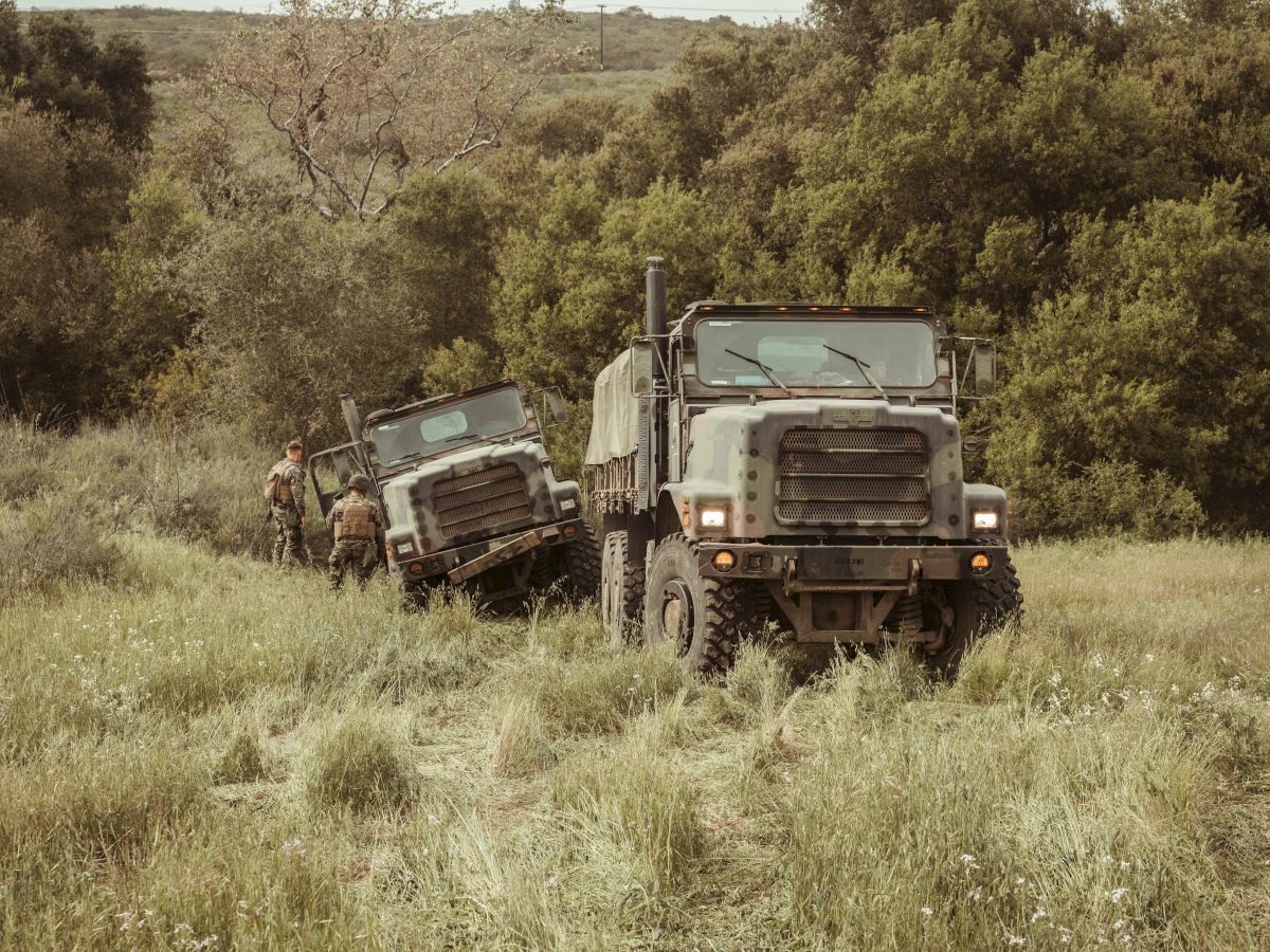 Two military trucks driving through a grassy field, with soldiers walking beside them in a wooded area.