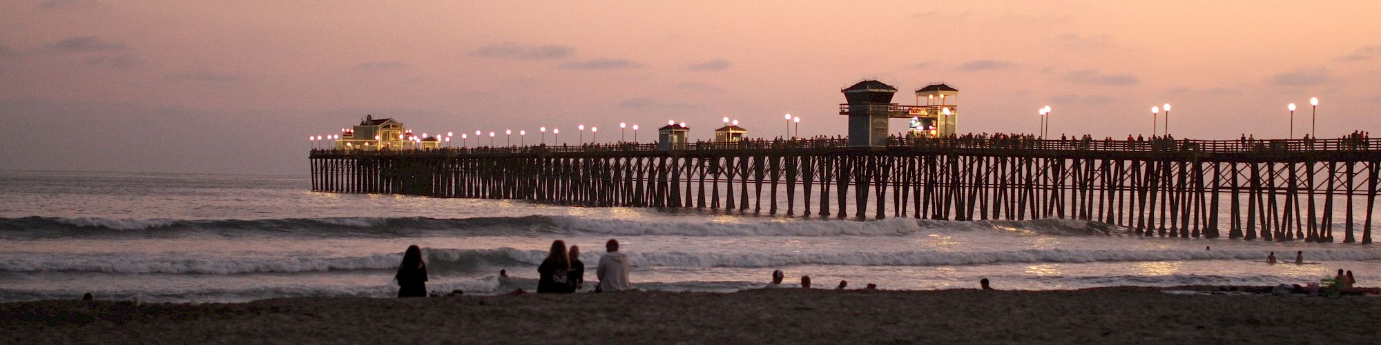 The image shows a beach at sunset with several people sitting on the sand, facing a lit pier extending over the ocean, under a pinkish sky.