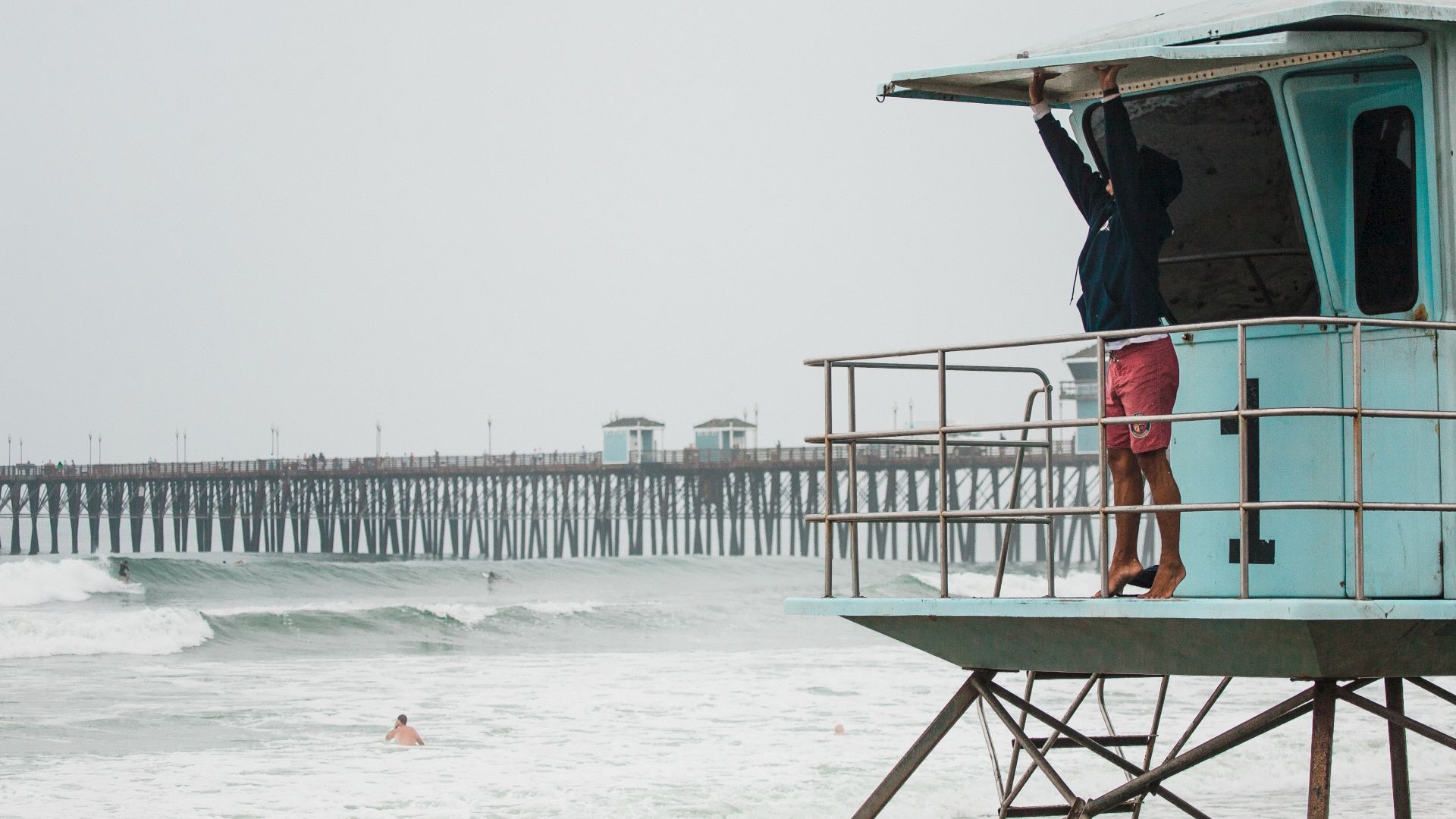 A person in pink shorts is standing on a lifeguard tower near the shore, looking out toward a pier extending into the ocean.