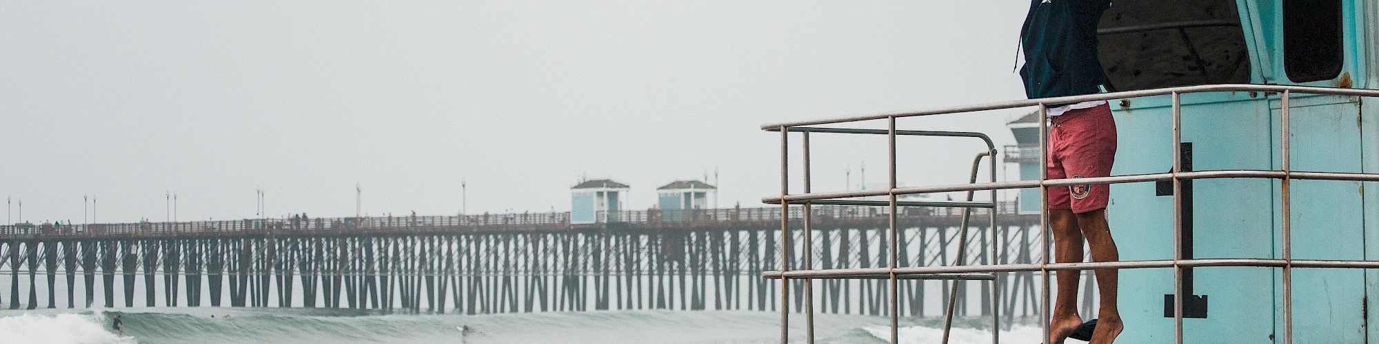 A person in pink shorts is standing on a lifeguard tower near the shore, looking out toward a pier extending into the ocean.