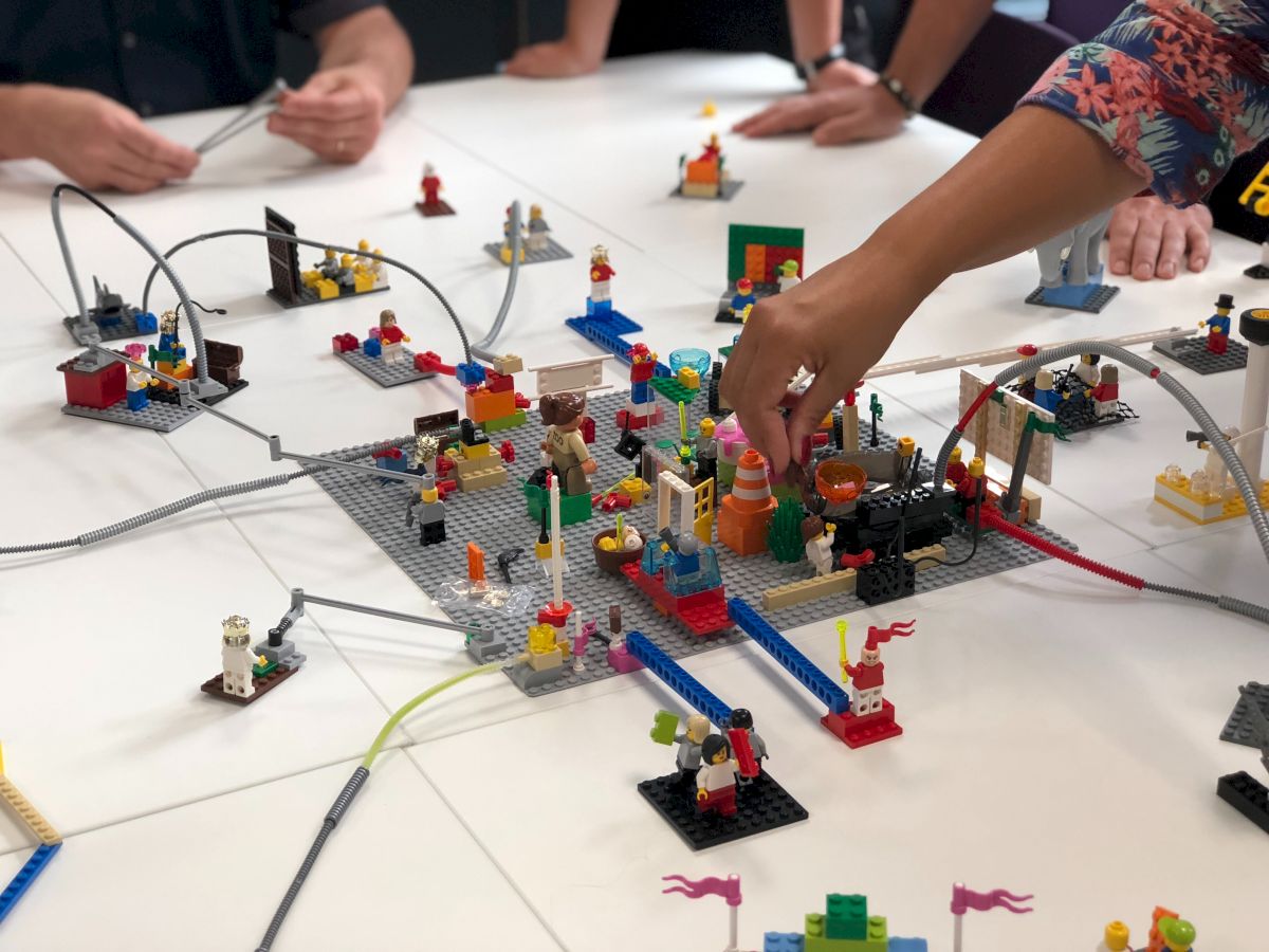 People are building and arranging LEGO pieces on a white table, creating a colorful and intricate layout.