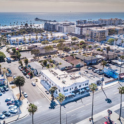 Aerial view of a coastal city with buildings, palm trees, parked cars, and the ocean in the background under a clear sky.