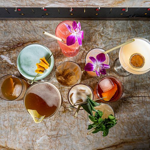 The image shows an assortment of colorful cocktails on a marble surface, decorated with flowers, citrus slices, and mint.