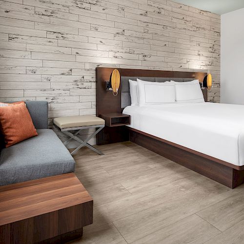 A modern hotel room with a large bed, wooden accents, a gray sofa with an orange pillow, a small table, a mirror, and light wood flooring.