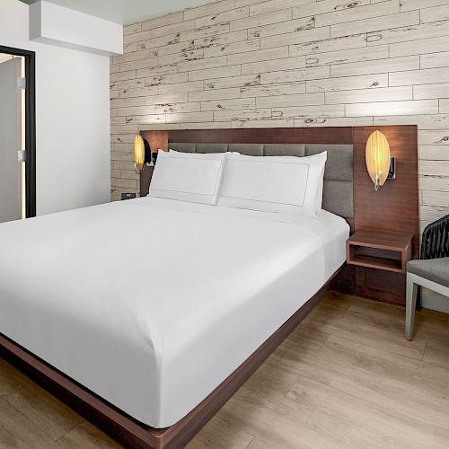 A neatly made bed in a modern hotel room, featuring wooden flooring, a cozy chair with a cushion, and a wood-paneled accent wall with soft lighting.