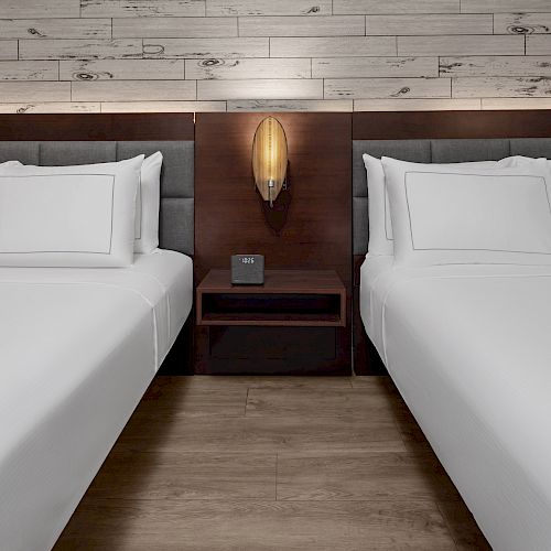 The image features two neatly made beds with white linens, a wooden headboard, a central nightstand, and a wall-mounted light fixture above.