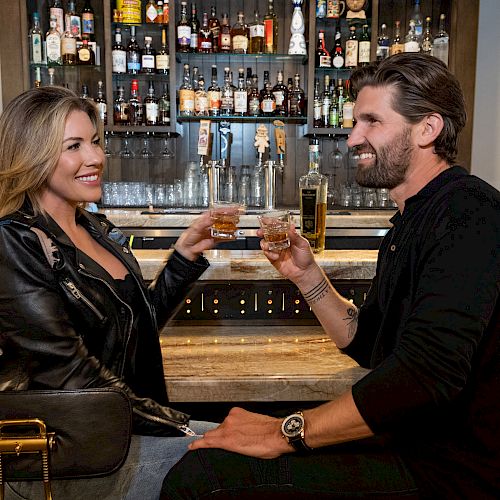 Two people are sitting at a bar, holding drinks and smiling at each other. The background shows a variety of liquor bottles.