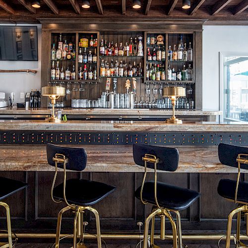 A stylish, well-lit bar with wooden finishes, black and gold stools, a fully stocked shelf, and golden lamps, situated next to large windows.