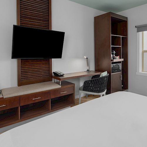 A hotel room with a TV, desk, chair, coffee maker, lamp, microwave, and a window is depicted in the image, seen from the foot of the bed.