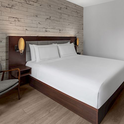 The image shows a modern hotel room with a neatly made bed, an accent wall, a wooden chair, and bedside lamps. The room has simple, elegant decor.