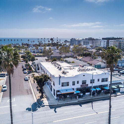 Aerial view of a coastal town with palm trees, buildings, parked cars, and the ocean in the background on a sunny day.